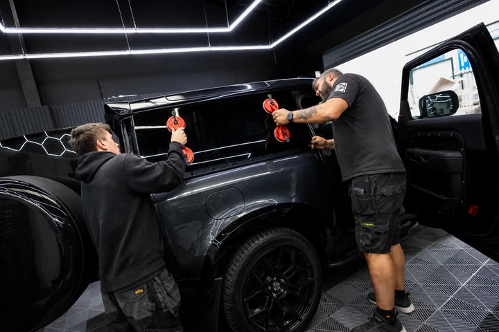 Two men adding a new replacement rear window into a grey land rover defender commercial to the rear side window of a black L663 SUV inside a modern auto shop with bright ceiling lights.