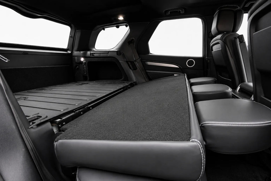 Rear cargo area of a black discovery 5 SUV L462 with the 2nd-row seat folded flat, showing a carpeted floor and the folded-up front seats against bright white window light.