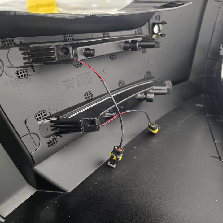 Close-up of an automotive electronic module showing black plastic mounting brackets, wiring harnesses, and connector plugs with red and black wires inside a vehicle compartment.
