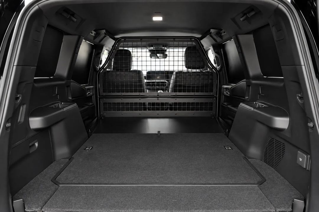 Rear cargo area of a black SUV with the tailgate open, showing a flat carpeted cargo bed, side pockets, and a rear mesh bulk head partition between the cargo area and the back seats.