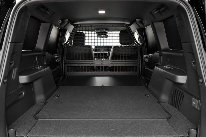 Rear cargo area of a black SUV with the tailgate open, showing a flat carpeted cargo bed, side pockets, and a rear mesh bulk head partition between the cargo area and the back seats.