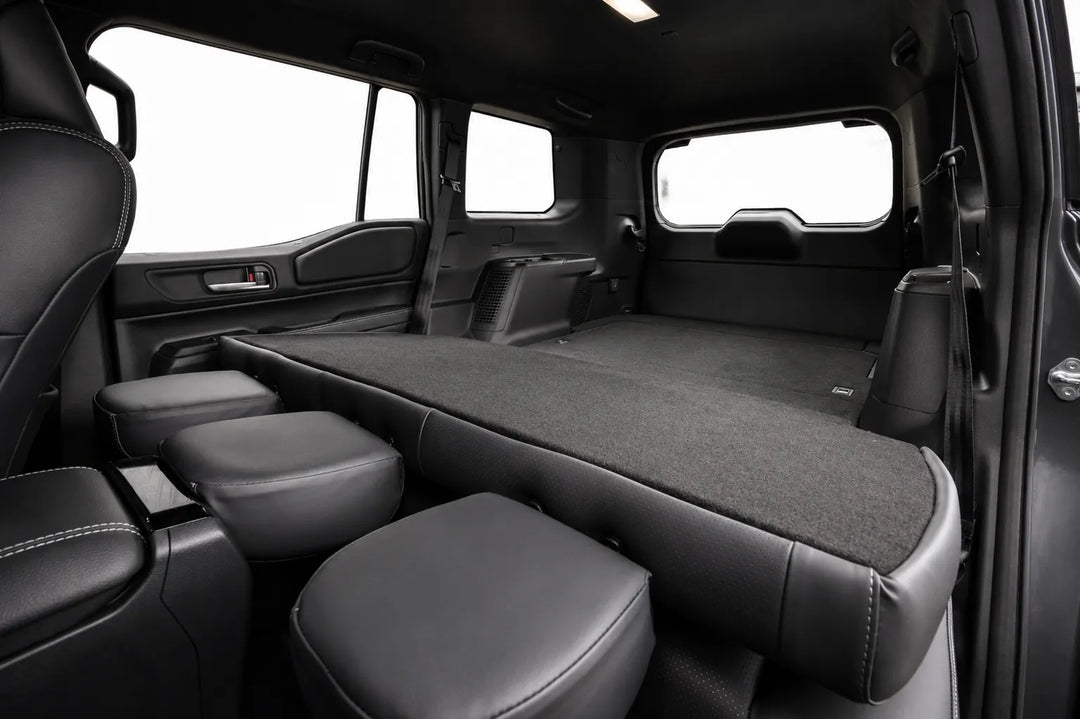 The interior cargo bed of a vehicle after the conversion showing the seats folded flat, featuring a dark gray carpeted floor and black leatherette seat bolsters and headrests, photographed from the side door opening.