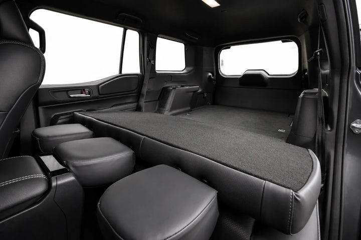 The interior cargo bed of a vehicle after the conversion showing the seats folded flat, featuring a dark gray carpeted floor and black leatherette seat bolsters and headrests, photographed from the side door opening.