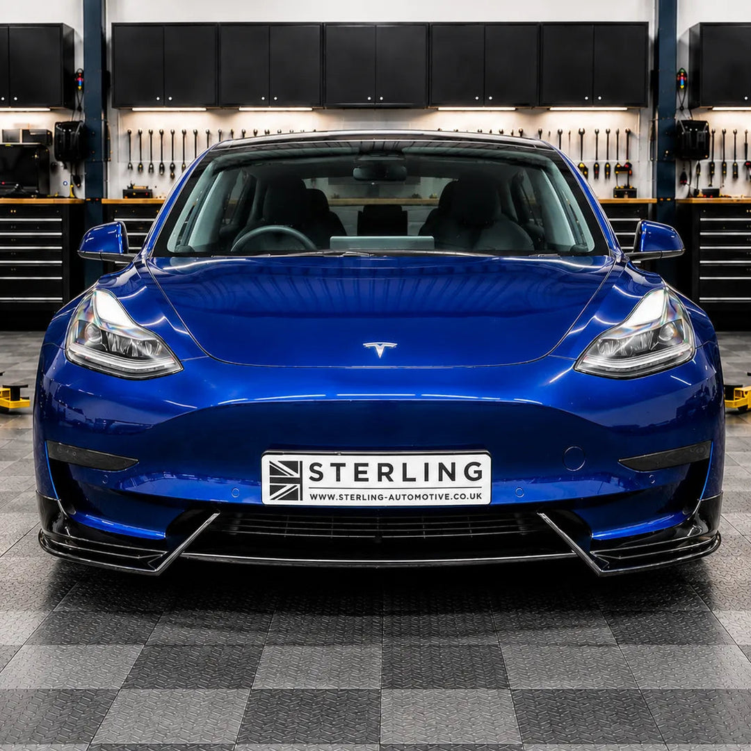 A blue Tesla Model 3 with a custom front splitter and a 'STERLING' license plate, photographed in the sterling garage setting with a checkered floor and a 'STERLING BRITISH AUTOMOTIVE DESIGN' sign on the wall behind it.