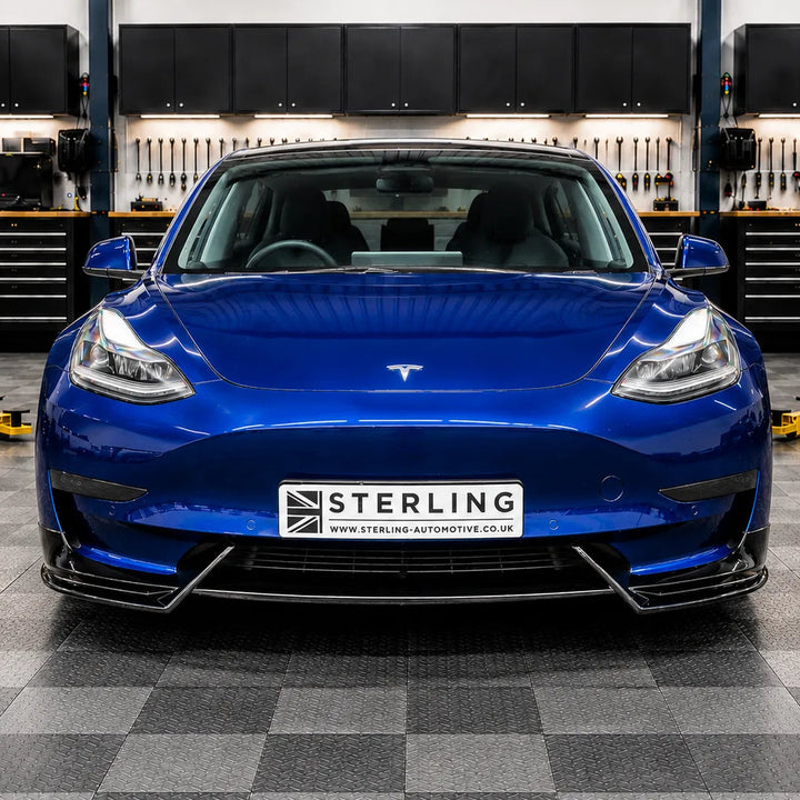 A blue Tesla Model 3 with a custom front splitter and a 'STERLING' license plate, photographed in the sterling garage setting with a checkered floor and a 'STERLING BRITISH AUTOMOTIVE DESIGN' sign on the wall behind it.