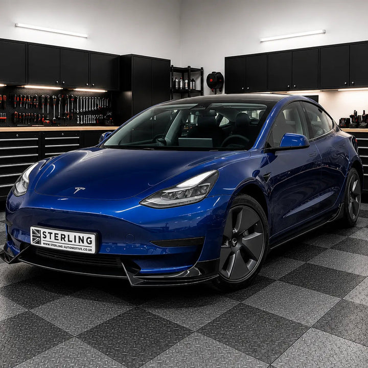 A metallic blue Tesla Model 3 parked inside a modern garage workshop with black tool cabinets and a gray checkered floor. A license plate reading 'STERLING' is mounted on the front.