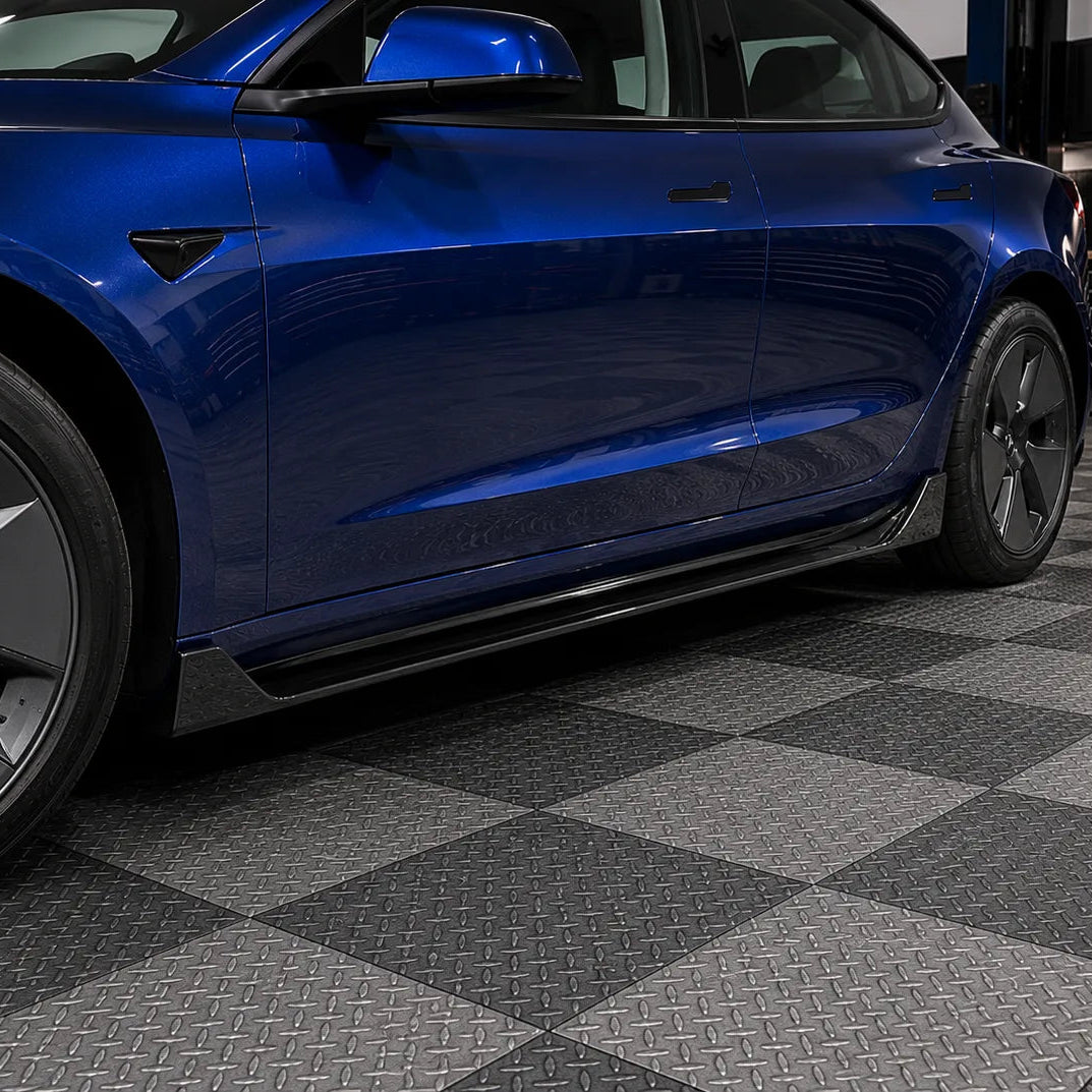 A close-up side view of a glossy blue Tesla Model 3 parked on a black and gray diamond-plate checkered floor inside a garage.