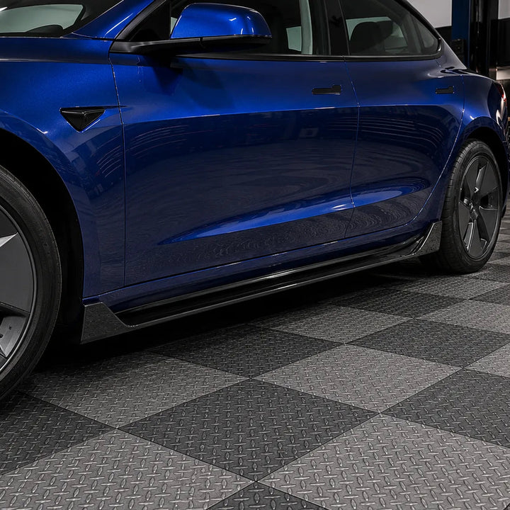 A close-up side view of a glossy blue Tesla Model 3 parked on a black and gray diamond-plate checkered floor inside a garage.