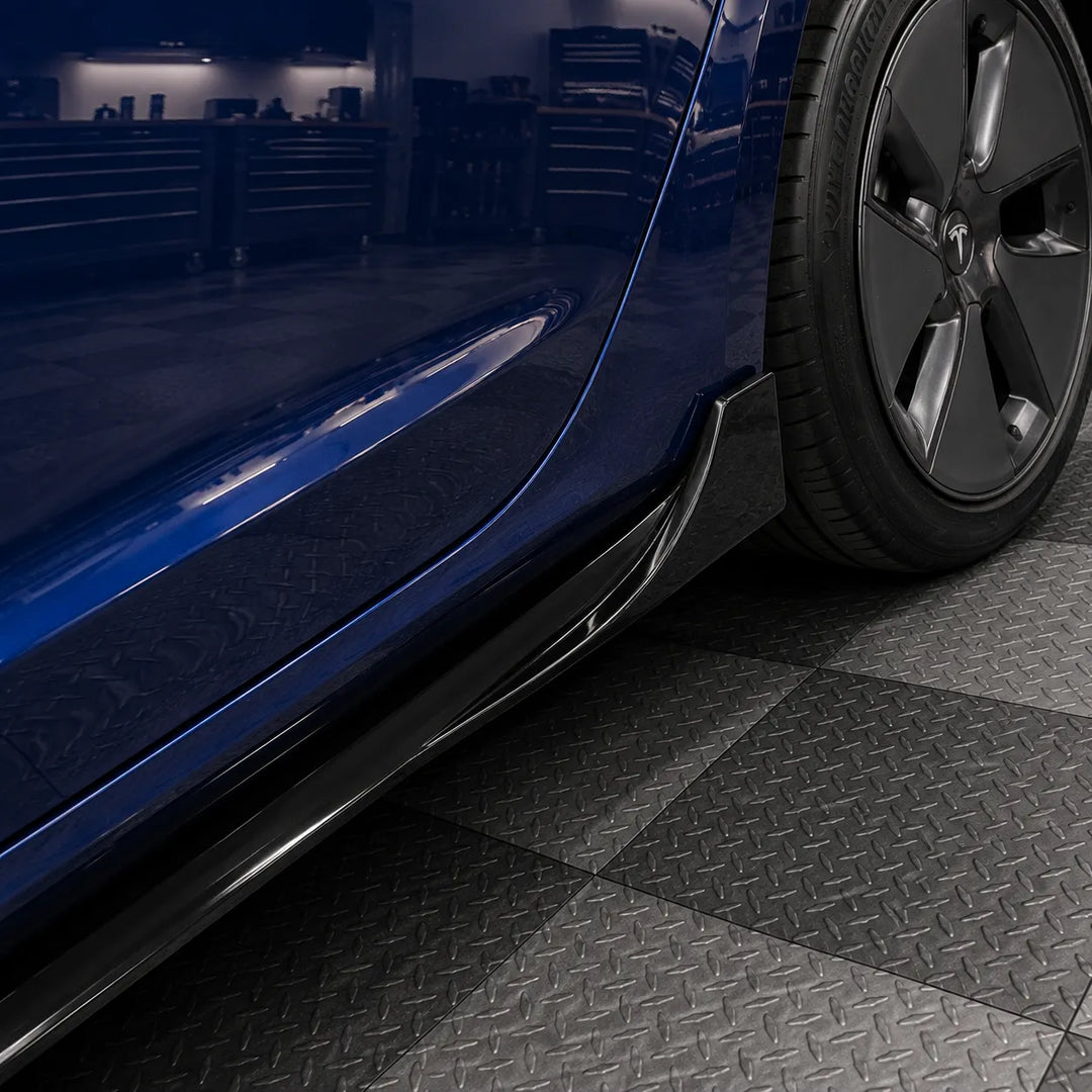 A close-up of a dark blue car's side body kit, including a glossy black side skirt and lip spoiler, mounted on a vehicle with a black Michelin tire, set against a dark garage background with diamond-plate flooring.