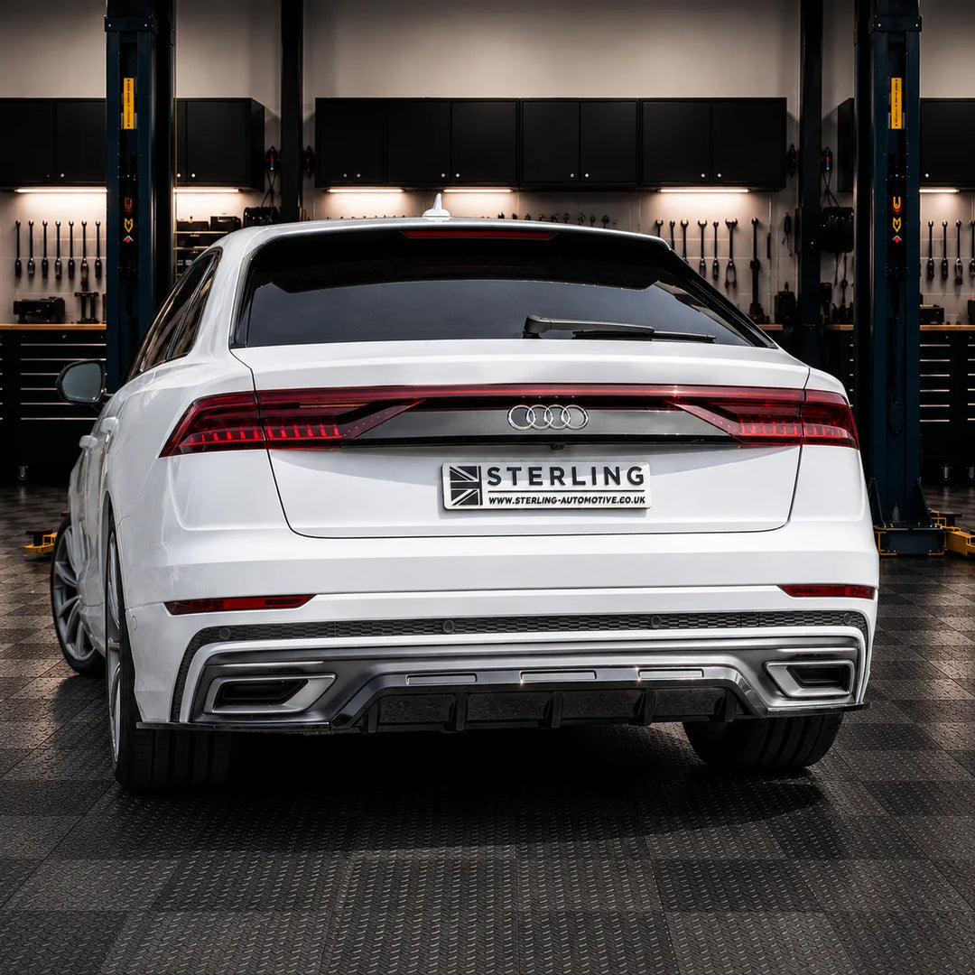 A white Audi sedan shown from the rear in a garage setting, with a Sterling Automotive badge mounted on the trunk. The car features a chrome diffuser, dual exhausts, and a clean, professional backdrop of a workshop.