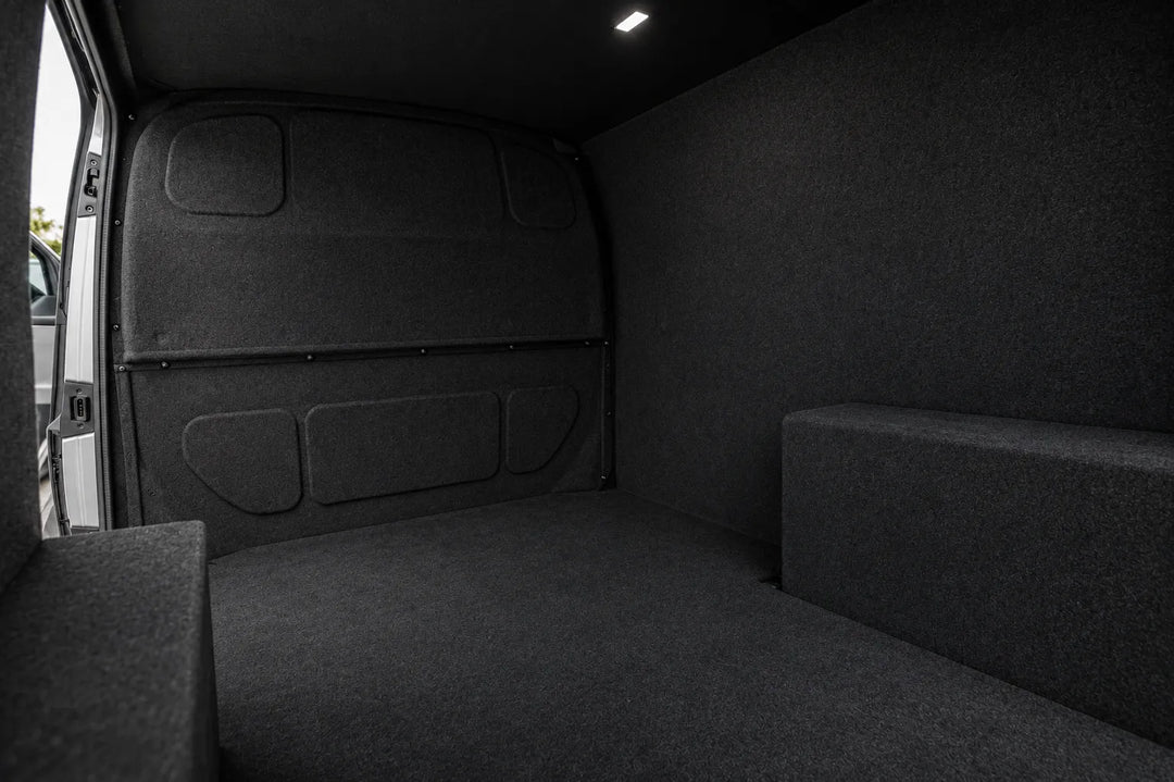 The interior cargo area of a vehicle with dark gray carpeted walls and floor, showing a raised side platform and a small overhead light.