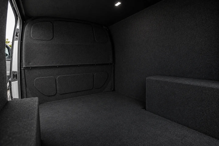 The interior cargo area of a vehicle with dark gray carpeted walls and floor, showing a raised side platform and a small overhead light.