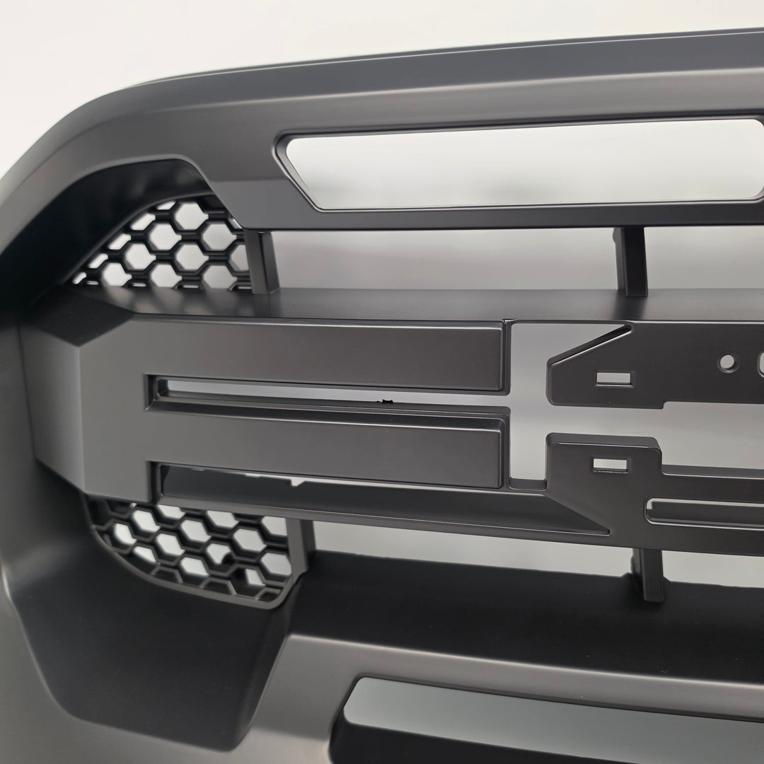 Close-up of a dark gray metallic component with geometric cutouts, honeycomb patterns, and mounting slots, photographed against a light neutral background.