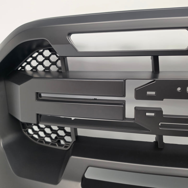 Close-up of a dark gray metallic component with geometric cutouts, honeycomb patterns, and mounting slots, photographed against a light neutral background.
