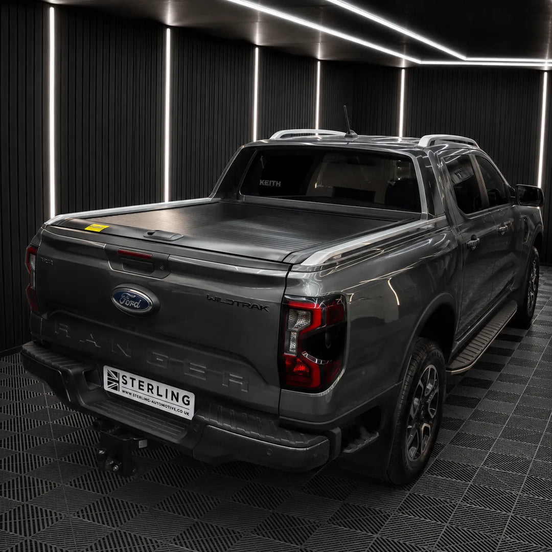 A dark gray Ford Ranger pickup truck shown from the rear three-quarter angle, fitted with a tonneau cover on the bed, parked inside a modern garage with black paneled walls and LED strip lighting.