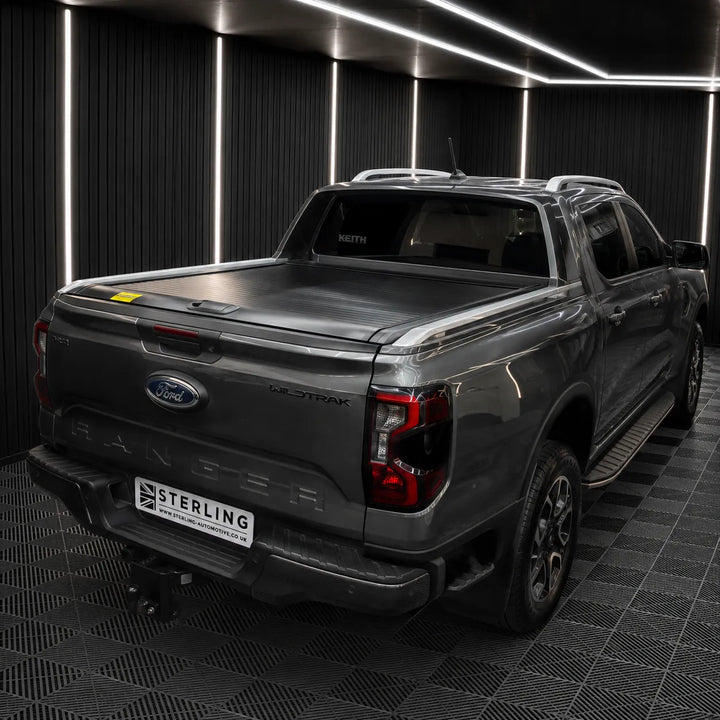 A dark gray Ford Ranger pickup truck shown from the rear three-quarter angle, fitted with a tonneau cover on the bed, parked inside a modern garage with black paneled walls and LED strip lighting.