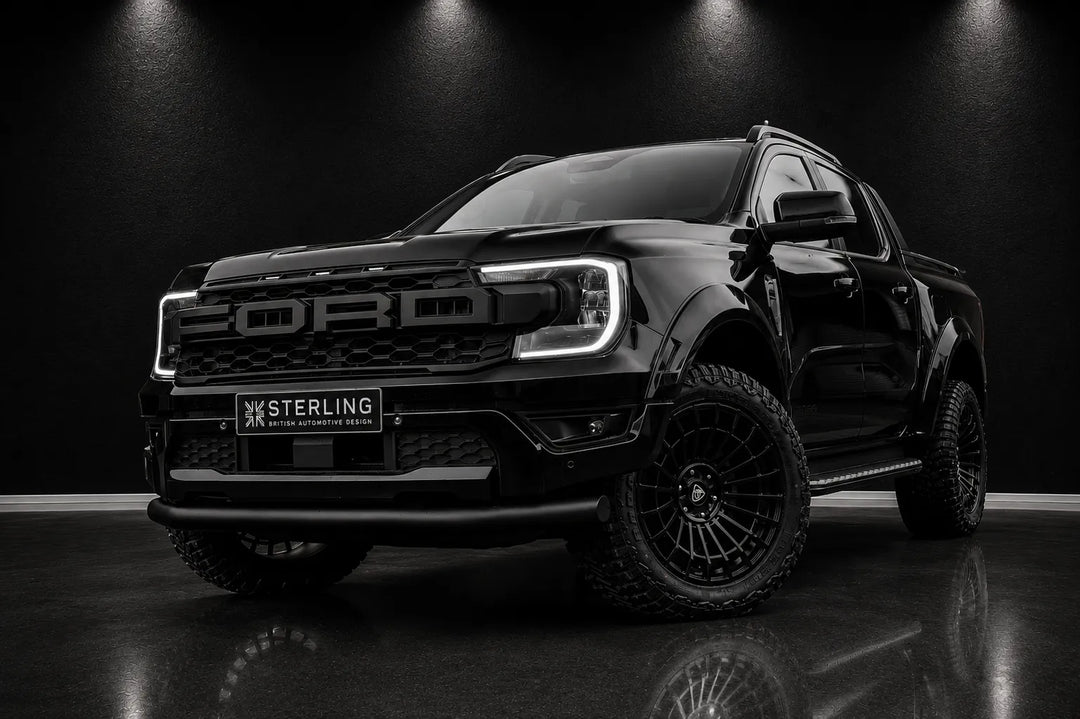A black Ford Ranger pickup truck with off-road tires and black multi-spoke wheels, parked on a dark reflective floor under spotlighting. A 'STERLING' license plate frame is mounted on the front bumper.