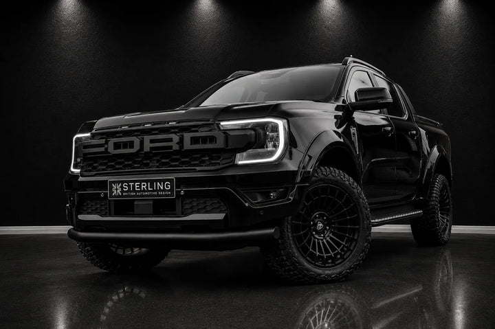 A black Ford Ranger pickup truck with off-road tires and black multi-spoke wheels, parked on a dark reflective floor under spotlighting. A 'STERLING' license plate frame is mounted on the front bumper.