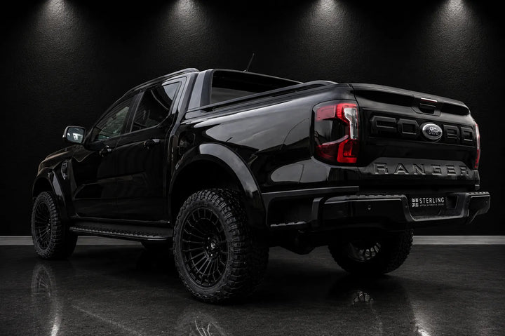 A black Ford Ranger pickup truck shown from a rear three-quarter angle in a dark studio setting with spotlights on a black floor.