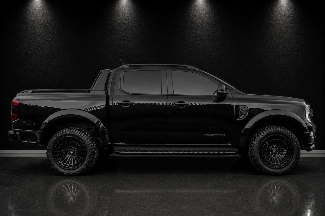 A black Ford Ranger pickup truck shown in a side profile view, parked on a reflective dark floor against a black background with overhead spotlighting.