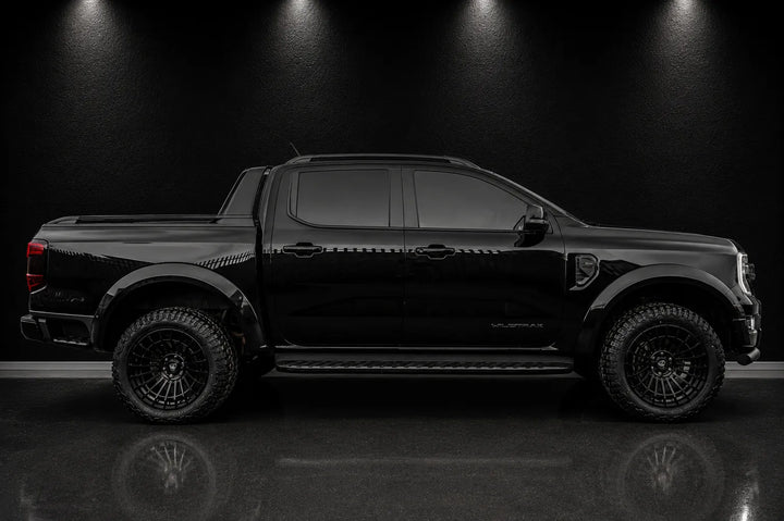 A black Ford Ranger pickup truck shown in a side profile view, parked on a reflective dark floor against a black background with overhead spotlighting.