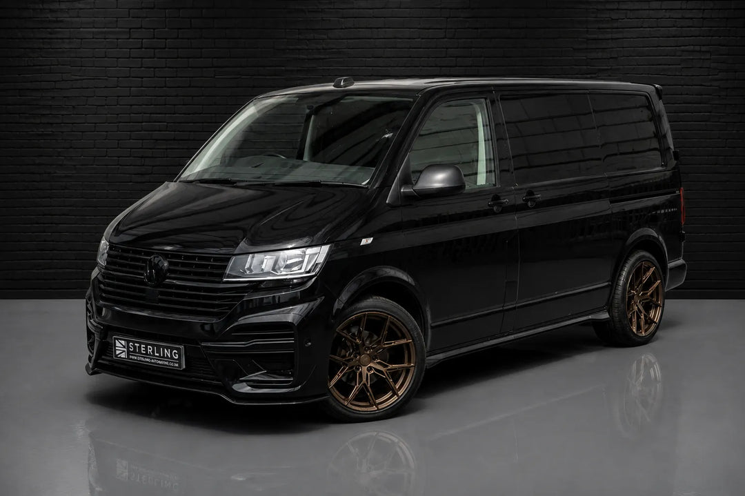 A black Volkswagen Transporter van with custom bronze multi-spoke wheels and a custom front bumper featuring Sterling Design Pack and a 'STERLING' badge, photographed in a studio setting against a dark brick wall.