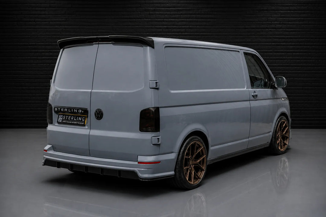 A light gray Volkswagen Transporter van with bronze-coloured alloy wheels, a rear spoiler, and a custom 'Sterling' branded rear badge, with the sterling design pack added, photographed in a studio setting against a dark brick wall.