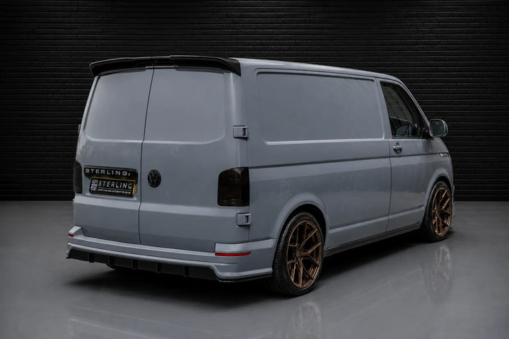 A light gray Volkswagen Transporter van with bronze-coloured alloy wheels, a rear spoiler, and a custom 'Sterling' branded rear badge, with the sterling design pack added, photographed in a studio setting against a dark brick wall.