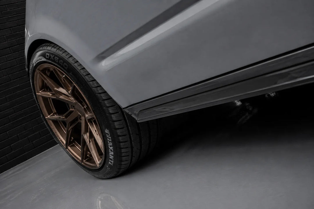 Close-up of a car wheel featuring a multi-spoke bronze alloy rim and a black side sill, mounted on a grey vehicle body in a studio setting.