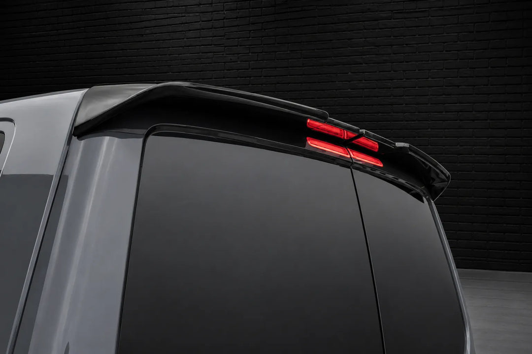 Close-up rear view of a silver van with a black roof spoiler and illuminated red taillights, set against a dark brick wall background.