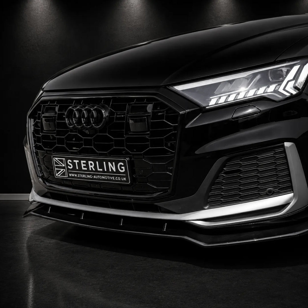 Close-up of the front grille and lower bumper area of a black Audi, showing a branded Sterling automotive grille insert and license plate frame. The car is photographed in a dark studio setting with dramatic lighting.