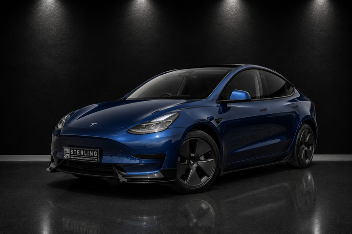 A dark metallic blue Tesla Model Y is shown from a front three-quarter angle in a studio setting with a Sterling Design Pack supplied in gloss black on a black background and dramatic overhead lighting. The car features a custom front splitter and a license plate frame with the brand name 'STERLING'.