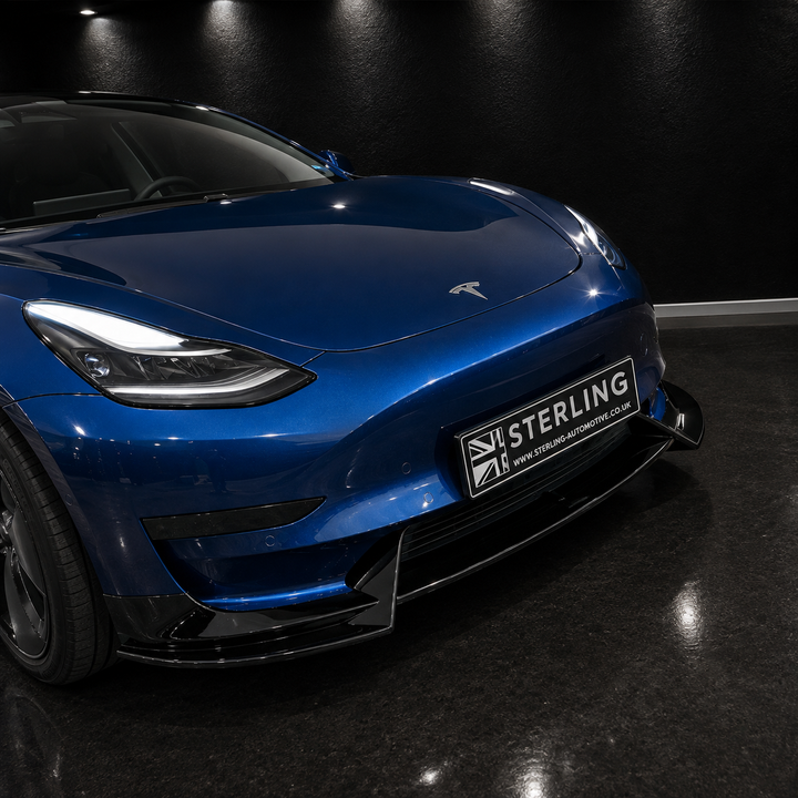 A blue Tesla Model Y shown from a low front three-quarter angle in a dark studio or showroom, with a black sterling front lip spoiler and a visible Sterling brand badge on the license plate area.
