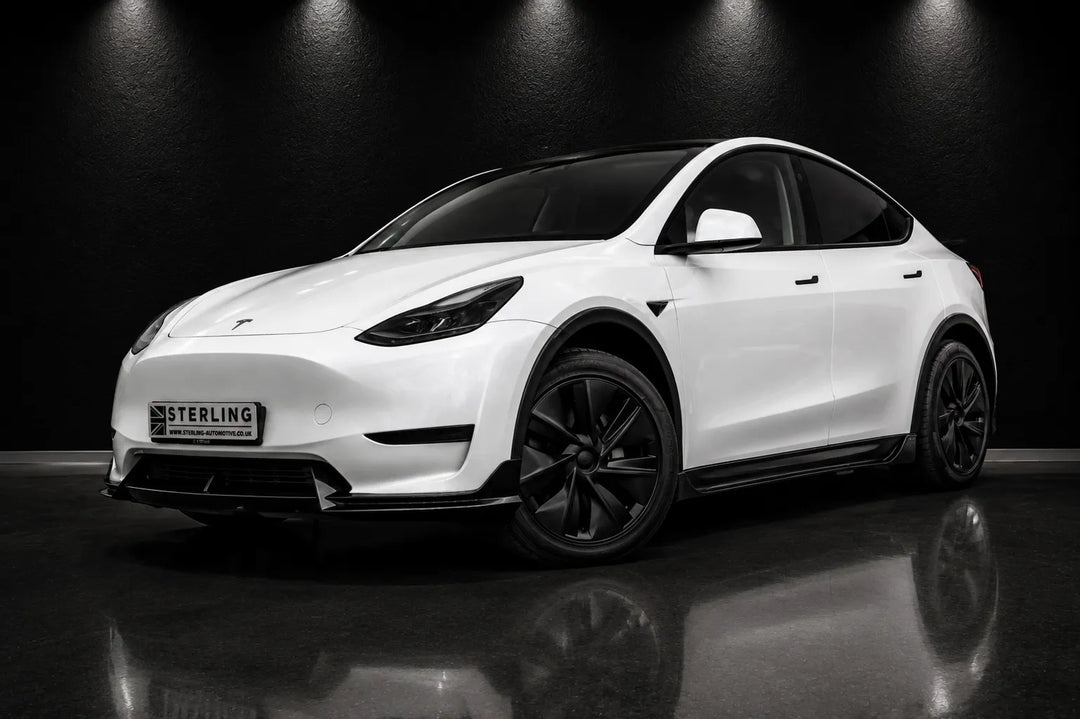 A white Tesla Model Y with black wheels and a black front lip kit, photographed in a dark studio setting with a reflection on the floor.