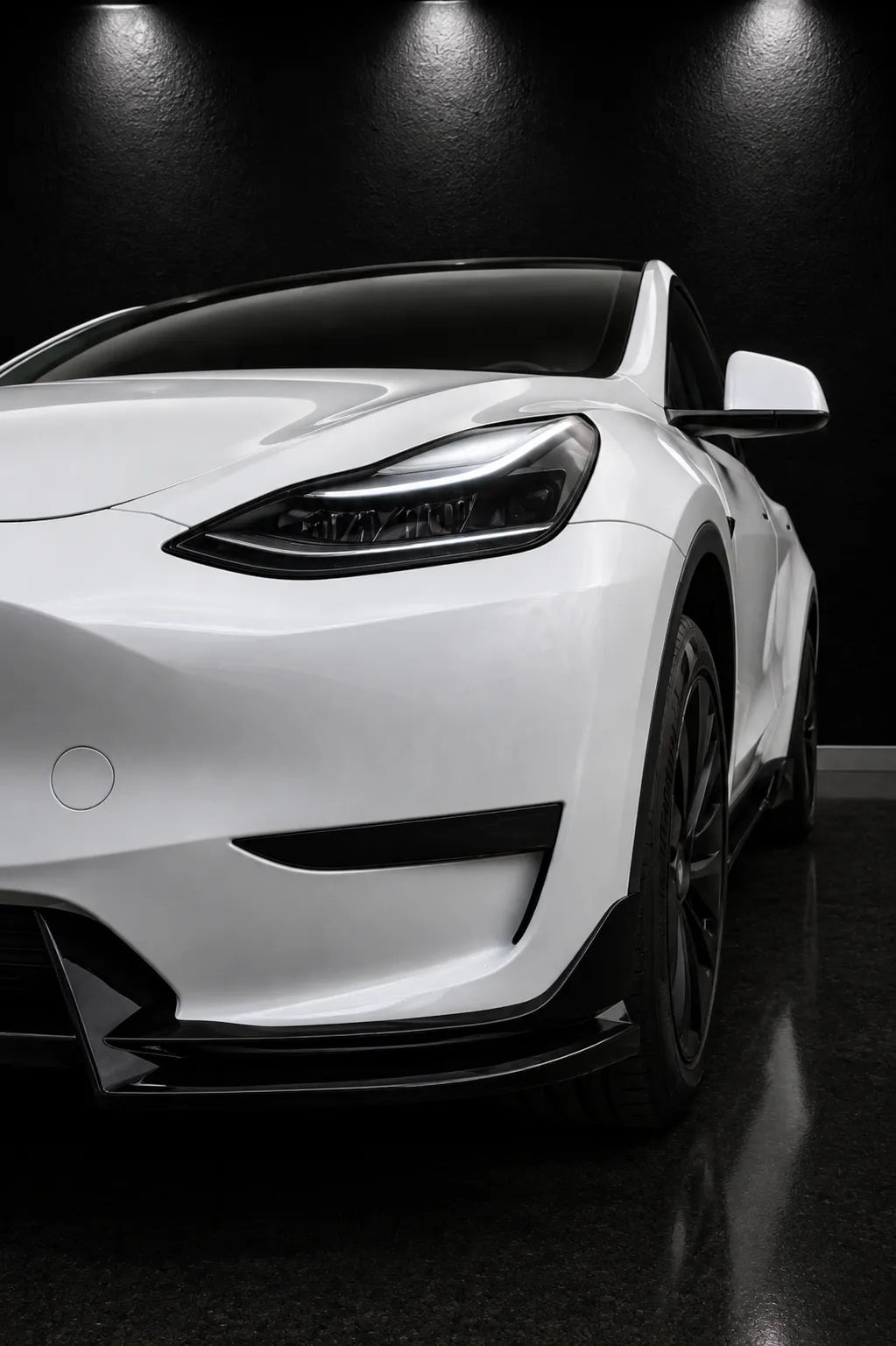 A white Tesla Model Y shown from a low front three-quarter angle in a dark studio setting with dramatic overhead lighting.