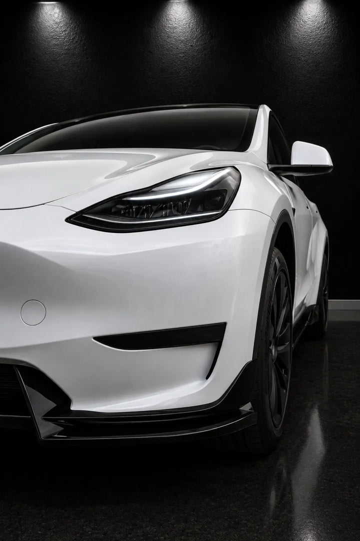 A white Tesla Model Y shown from a low front three-quarter angle in a dark studio setting with dramatic overhead lighting.