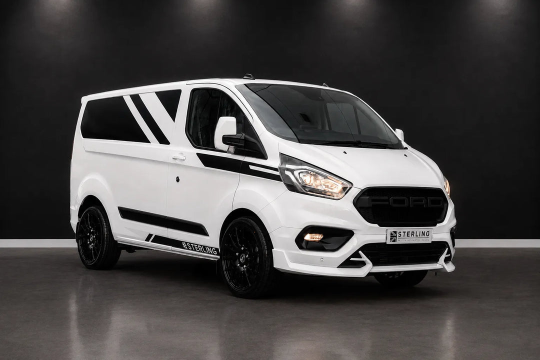 A white Ford Transit Custom van with custom black graphics, black wheels, and a custom front bumper by Sterling, photographed in a dark studio setting.