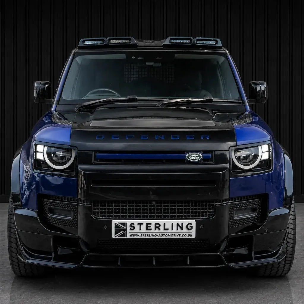 A front-facing view of a Boston Blue Land Rover Defender with a black widebody kit by Sterling, featuring a black front grille, lip spoiler, and roof-mounted LED Sterling light bars, set against a dark corrugated metal wall.