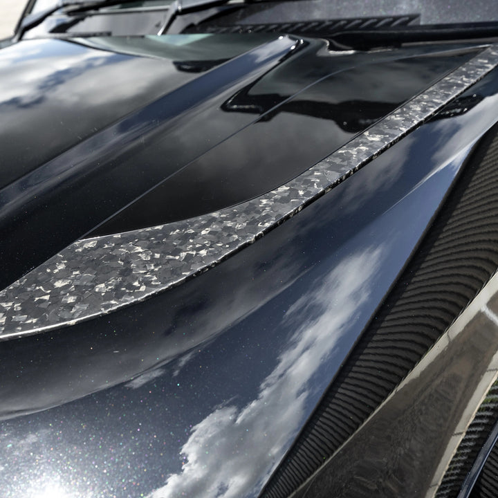 A close-up of a glossy black car hood featuring a Forged carbon fiber decorative accent strip running diagonally across the front.