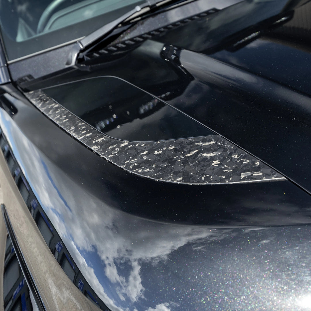 A close-up of a Forged carbon, glossy black front bonnet vent pair fitted on a black land rover defender L663 car hood with a textured clear carbon fiber accent strip running across the front.