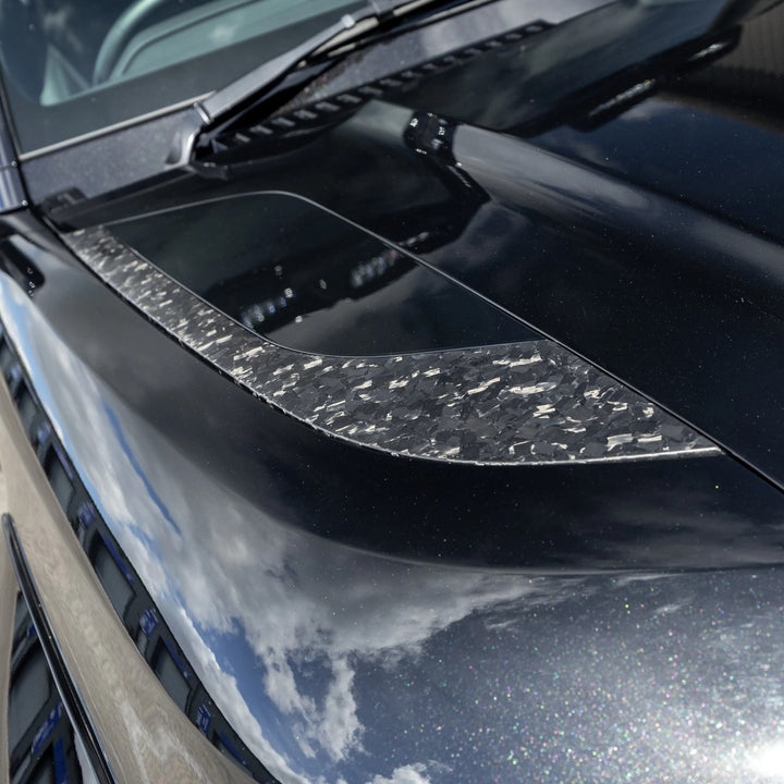 A close-up of a Forged carbon, glossy black front bonnet vent pair fitted on a black land rover defender L663 car hood with a textured clear carbon fiber accent strip running across the front.