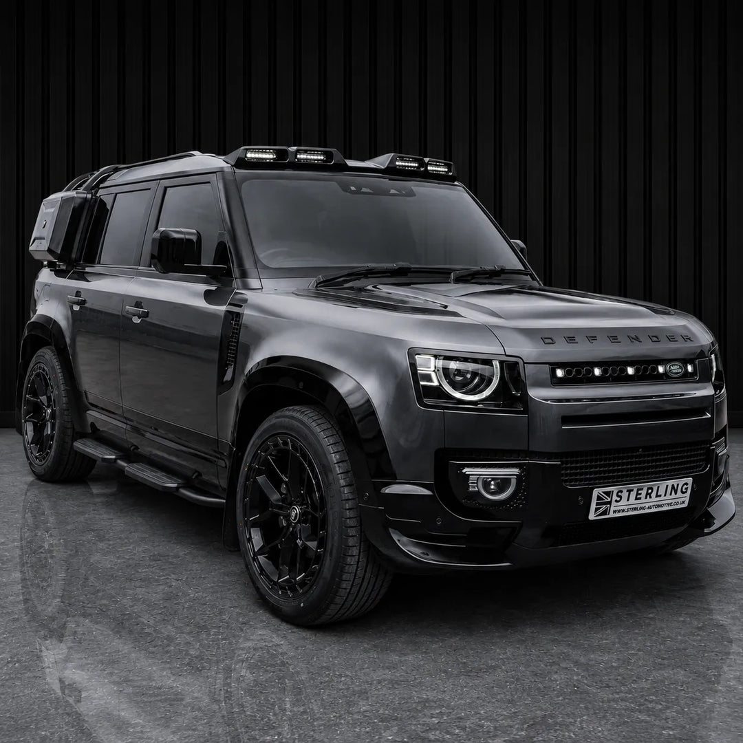 A black customized Defender SUV with a roof-mounted light bar, black wheels, and a custom front grille, parked against a dark vertical slat wall.