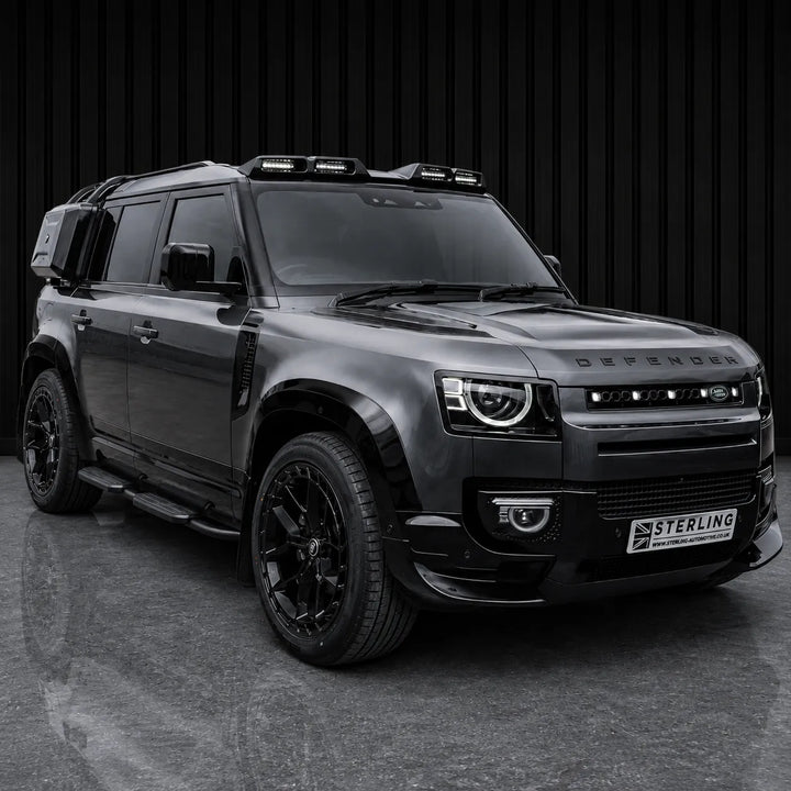 A black customized Defender SUV with a roof-mounted light bar, black wheels, and a custom front grille, parked against a dark vertical slat wall.